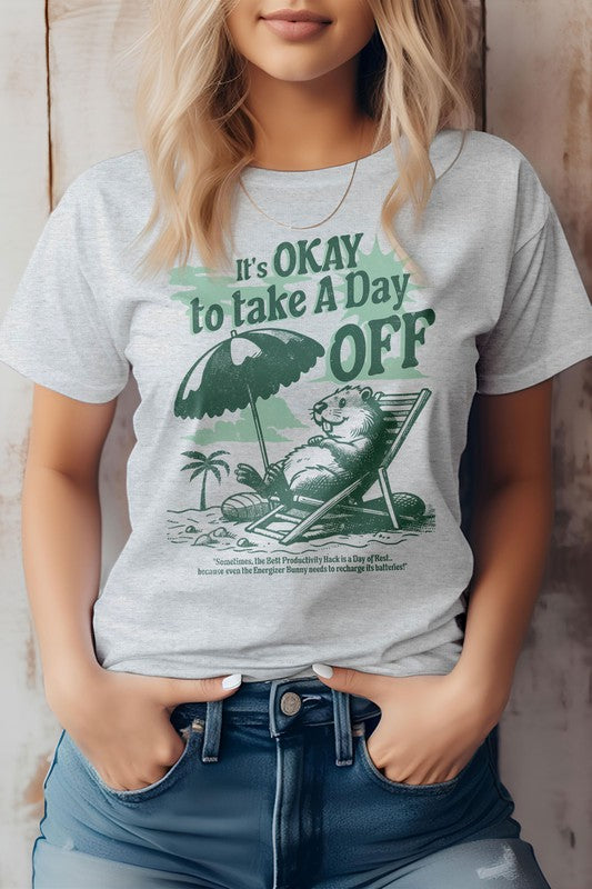 A person wearing the Day Off, Retro Vintage Graphic Tee in light green, featuring an illustration of a beaver relaxing on a beach under an umbrella. Made from eco-friendly materials, this shirt proudly displays the message "It's OKAY to Take A Day OFF" in bold letters.