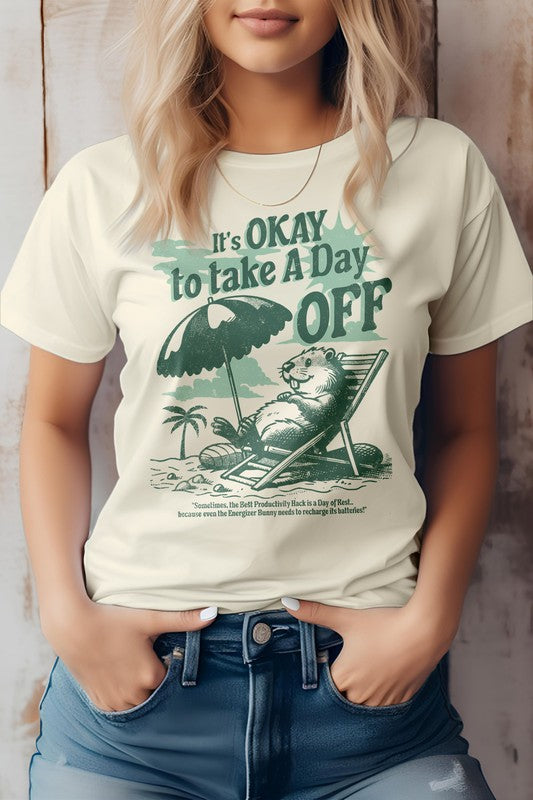 A person wearing the Day Off, Retro Vintage Graphic Tee in light green, featuring an illustration of a beaver relaxing on a beach under an umbrella. Made from eco-friendly materials, this shirt proudly displays the message "It's OKAY to Take A Day OFF" in bold letters.