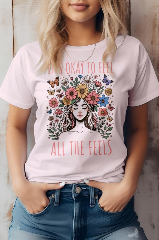 A person wearing the "It's Okay to Feel, All the Feels, Boho Graphic Tee," a light pink shirt featuring a woman surrounded by flowers and butterflies, with the text "It's okay to feel all the feels.