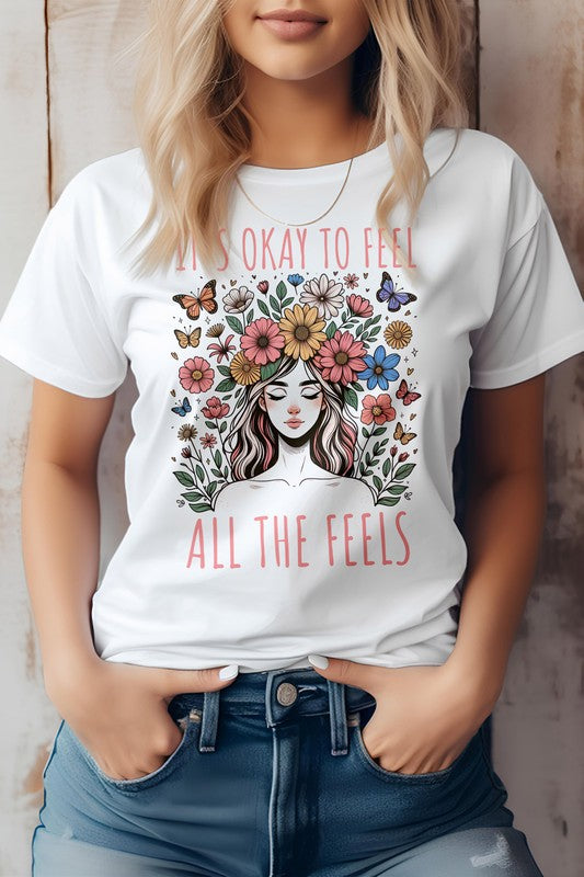 A person wearing the "It's Okay to Feel, All the Feels, Boho Graphic Tee," a light pink shirt featuring a woman surrounded by flowers and butterflies, with the text "It's okay to feel all the feels.