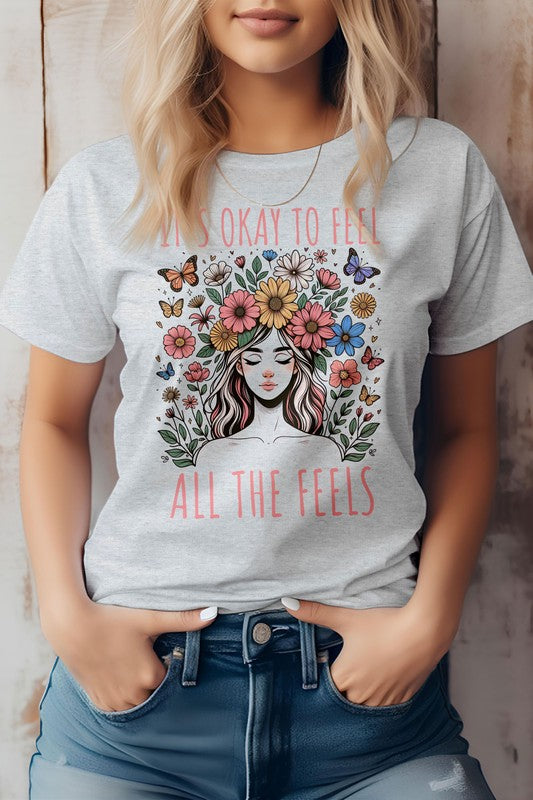 A person wearing the "It's Okay to Feel, All the Feels, Boho Graphic Tee," a light pink shirt featuring a woman surrounded by flowers and butterflies, with the text "It's okay to feel all the feels.