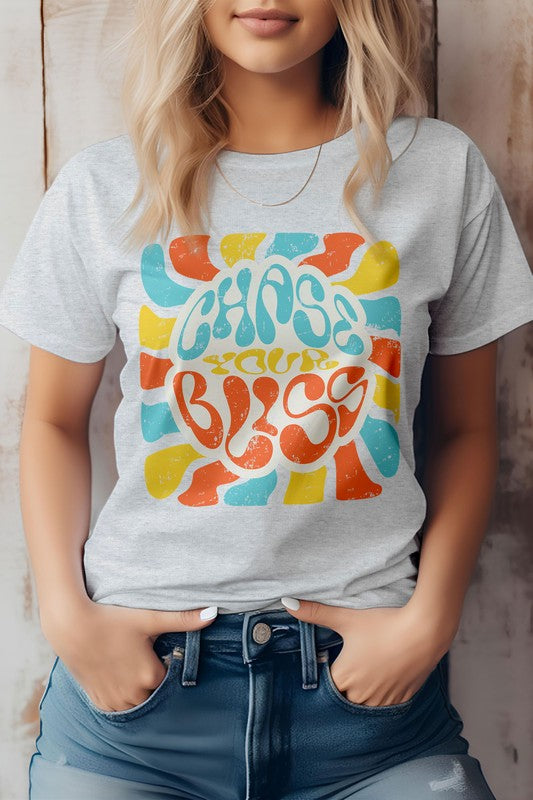 A person wearing a cream-colored "Chase Your Bliss, Retro Graphic Tee" with a colorful design stands in front of a wooden background, one hand casually tucked into their jeans pocket.