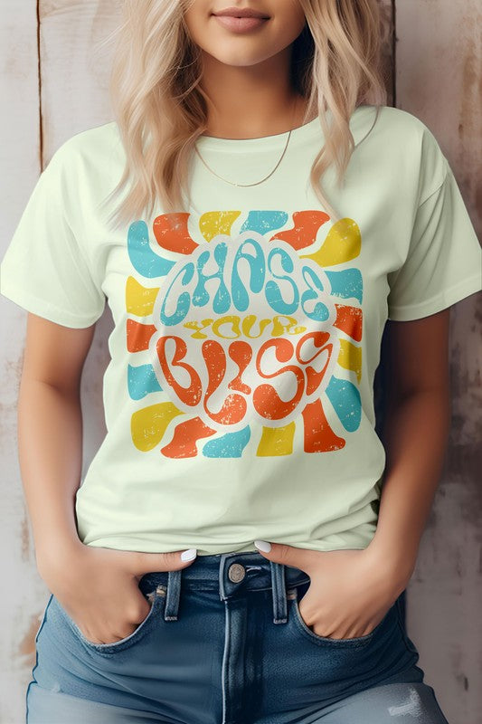 A person wearing a cream-colored "Chase Your Bliss, Retro Graphic Tee" with a colorful design stands in front of a wooden background, one hand casually tucked into their jeans pocket.