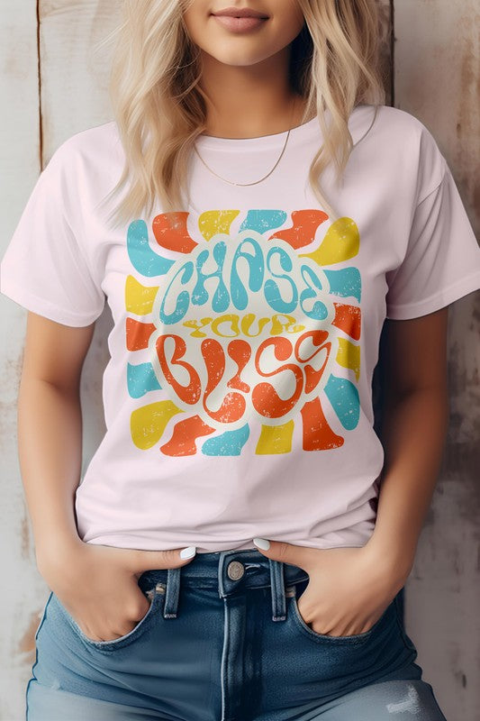 A person wearing a cream-colored "Chase Your Bliss, Retro Graphic Tee" with a colorful design stands in front of a wooden background, one hand casually tucked into their jeans pocket.