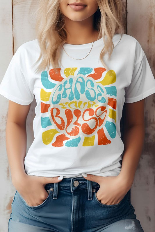 A person wearing a cream-colored "Chase Your Bliss, Retro Graphic Tee" with a colorful design stands in front of a wooden background, one hand casually tucked into their jeans pocket.