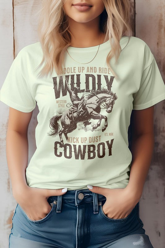 A person stands wearing the Kick up Dust Cowboy, Vintage Western Graphic Tee featuring the text "Saddle Up and Ride Wildly, Kick Up Dust, Cowboy" along with a graphic of a cowboy on horseback. This eco-friendly tee pairs perfectly with their hands in their jean pockets for an effortlessly cool look.