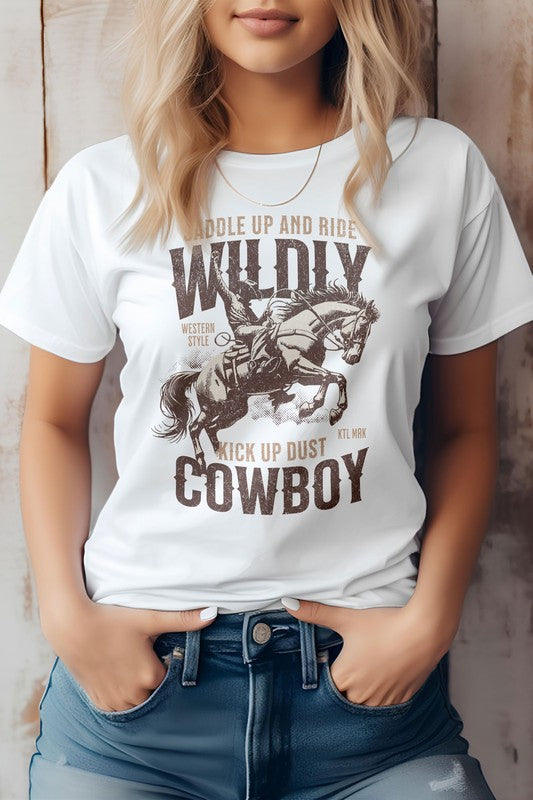 A person stands wearing the Kick up Dust Cowboy, Vintage Western Graphic Tee featuring the text "Saddle Up and Ride Wildly, Kick Up Dust, Cowboy" along with a graphic of a cowboy on horseback. This eco-friendly tee pairs perfectly with their hands in their jean pockets for an effortlessly cool look.