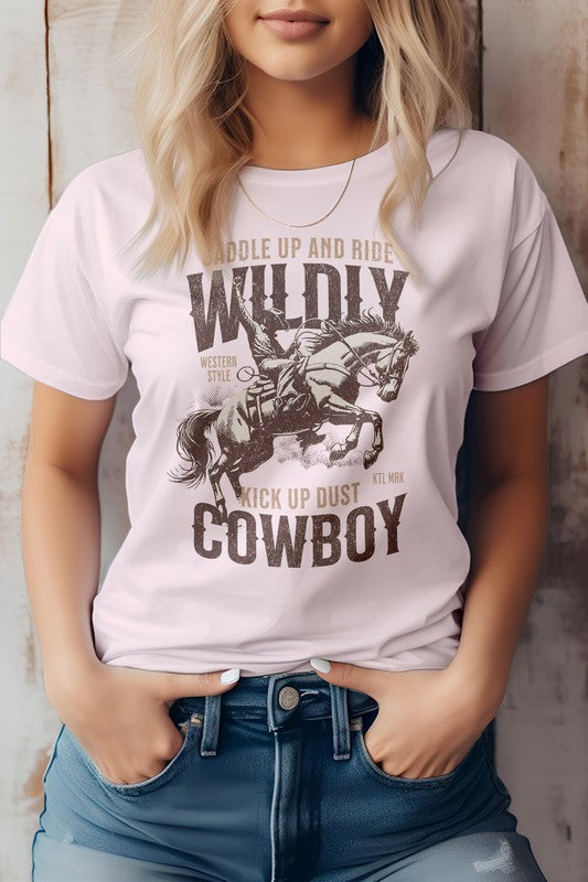 A person stands wearing the Kick up Dust Cowboy, Vintage Western Graphic Tee featuring the text "Saddle Up and Ride Wildly, Kick Up Dust, Cowboy" along with a graphic of a cowboy on horseback. This eco-friendly tee pairs perfectly with their hands in their jean pockets for an effortlessly cool look.