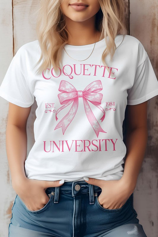 A person is wearing the Vintage Coquette University Graphic Tee made from combed and ring-spun cotton, featuring the text "Coquette University" and a bow graphic along with "Est. 1984." The individual stands with hands in the pockets of blue jeans.