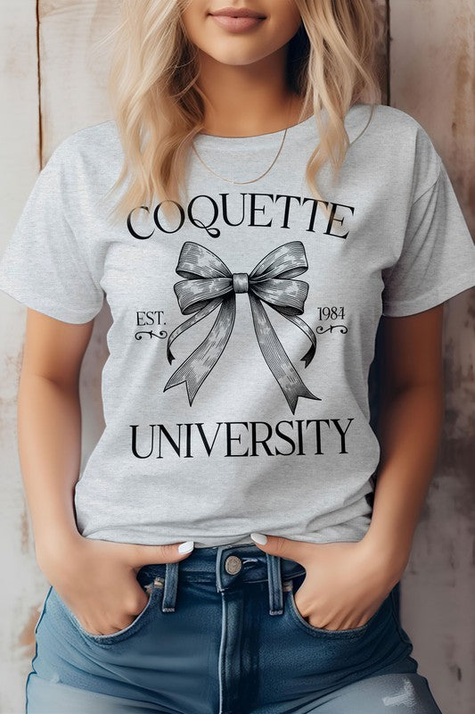 A person is wearing the Vintage Coquette University Graphic Tee made from combed and ring-spun cotton, featuring the text "Coquette University" and a bow graphic along with "Est. 1984." The individual stands with hands in the pockets of blue jeans.