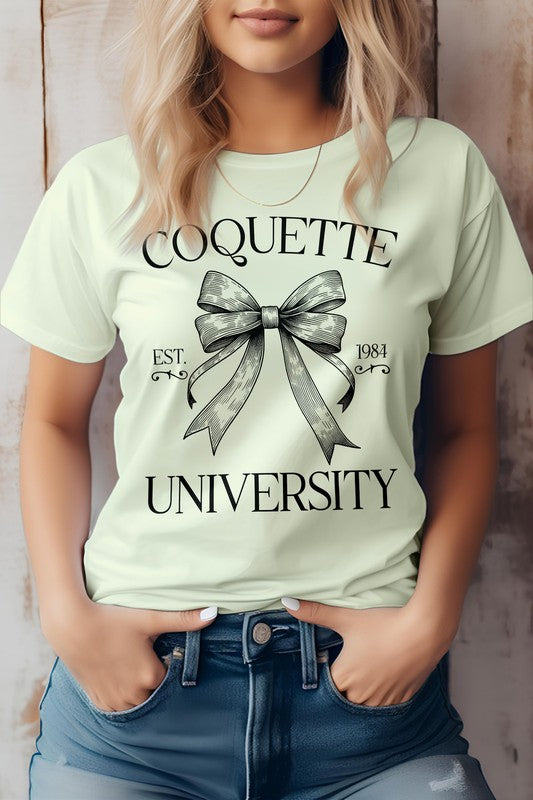 A person is wearing the Vintage Coquette University Graphic Tee made from combed and ring-spun cotton, featuring the text "Coquette University" and a bow graphic along with "Est. 1984." The individual stands with hands in the pockets of blue jeans.