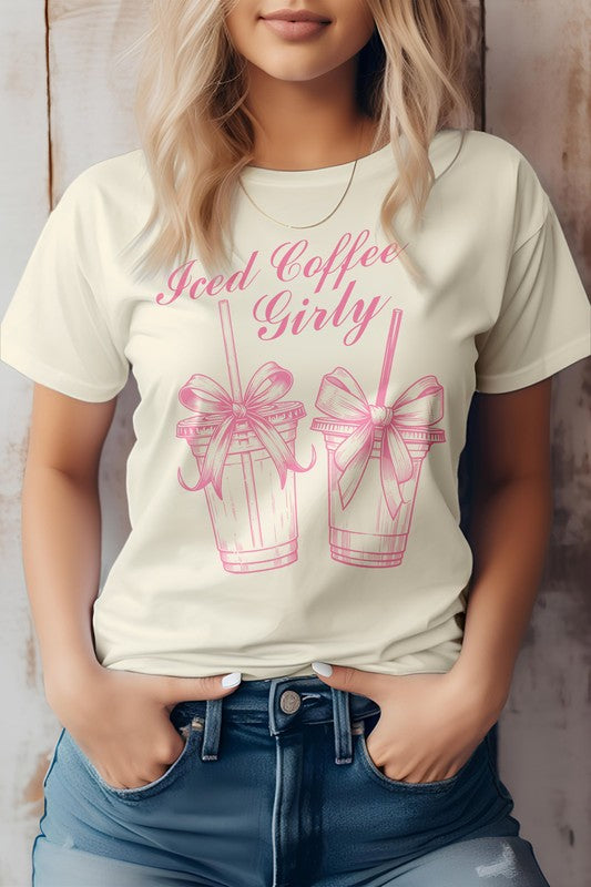 A woman wearing a Vintage Coquette Coffee Graphic Tee, made from eco-friendly materials, poses with her hands in her jean pockets. Her gray t-shirt features the text "Iced Coffee Girly" and graphic images of two iced coffee cups with bows.