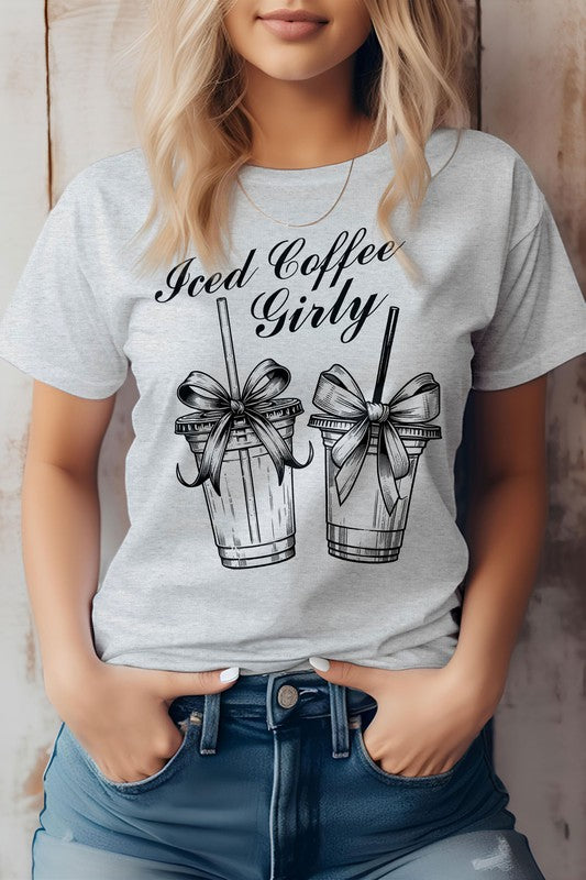 A woman wearing a Vintage Coquette Coffee Graphic Tee, made from eco-friendly materials, poses with her hands in her jean pockets. Her gray t-shirt features the text "Iced Coffee Girly" and graphic images of two iced coffee cups with bows.