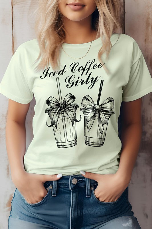 A woman wearing a Vintage Coquette Coffee Graphic Tee, made from eco-friendly materials, poses with her hands in her jean pockets. Her gray t-shirt features the text "Iced Coffee Girly" and graphic images of two iced coffee cups with bows.