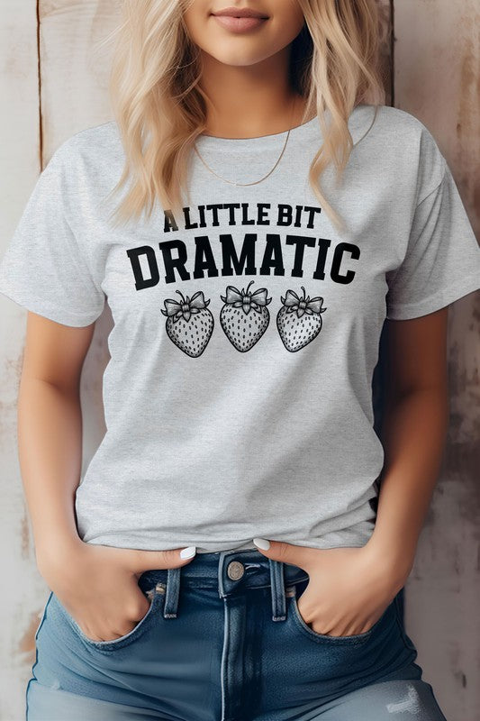 Person wearing a Vintage Coquette Strawberry Graphic Tee, featuring the text "A Little Bit Dramatic" and three illustrated strawberries below it, stands with hands in jean pockets. This stylish Bella Canvas tee is crafted from eco-friendly materials for a sustainable fashion choice.