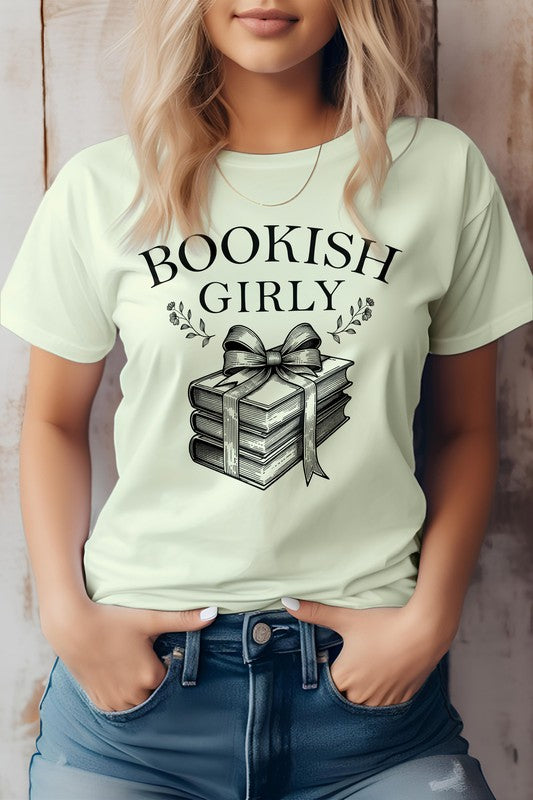 A person wearing a light-colored Vintage Coquette Book Graphic Tee made in the USA, featuring a stack of books tied with a ribbon. Crafted from eco-friendly materials, the person is shown from the shoulders down, hands in pockets.