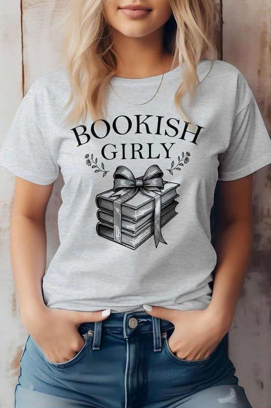 A person wearing a light-colored Vintage Coquette Book Graphic Tee made in the USA, featuring a stack of books tied with a ribbon. Crafted from eco-friendly materials, the person is shown from the shoulders down, hands in pockets.