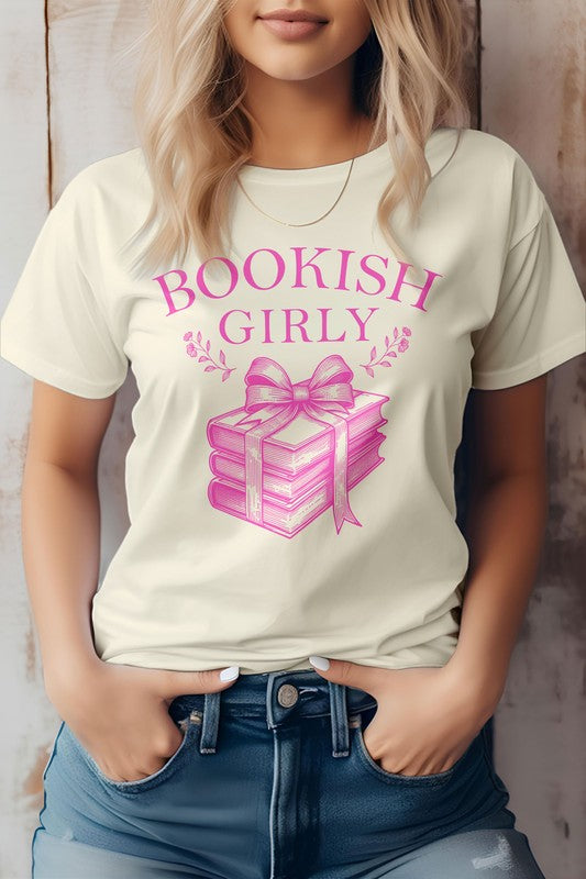 A person wearing a light-colored Vintage Coquette Book Graphic Tee made in the USA, featuring a stack of books tied with a ribbon. Crafted from eco-friendly materials, the person is shown from the shoulders down, hands in pockets.