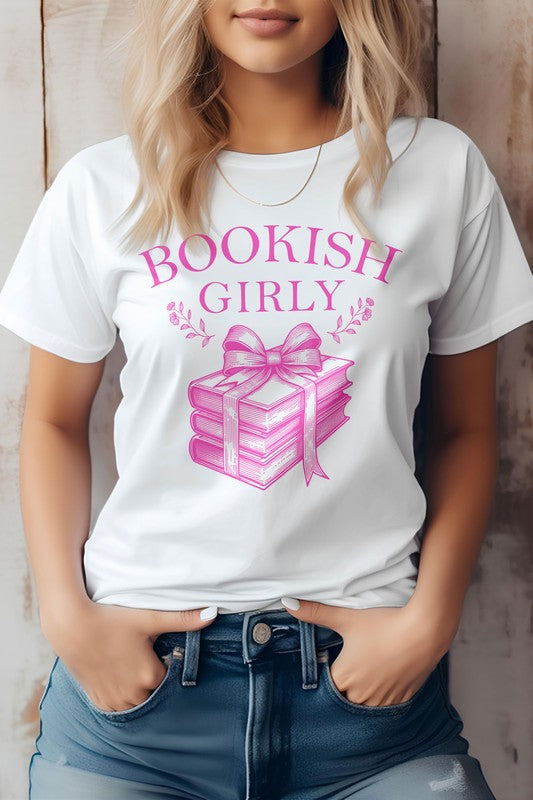 A person wearing a light-colored Vintage Coquette Book Graphic Tee made in the USA, featuring a stack of books tied with a ribbon. Crafted from eco-friendly materials, the person is shown from the shoulders down, hands in pockets.