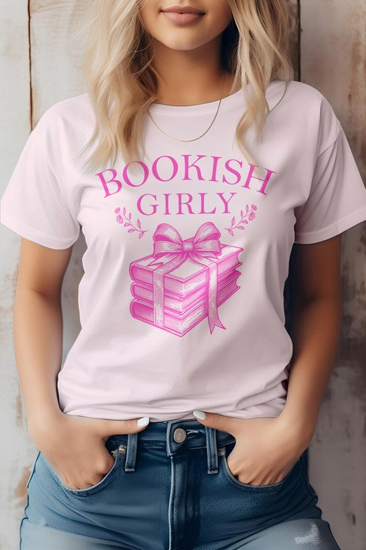 A person wearing a light-colored Vintage Coquette Book Graphic Tee made in the USA, featuring a stack of books tied with a ribbon. Crafted from eco-friendly materials, the person is shown from the shoulders down, hands in pockets.