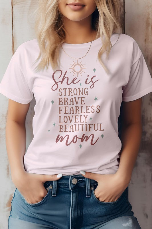 A woman wearing a light pink "She is Mom Graphic Tee" with text that reads, "She is strong, brave, fearless, lovely, beautiful mom." She has blonde hair and her hands are in her jean pockets—a perfect Mother's Day tee.