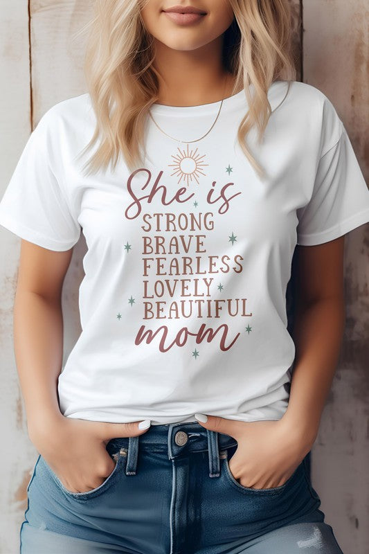 A woman wearing a light pink "She is Mom Graphic Tee" with text that reads, "She is strong, brave, fearless, lovely, beautiful mom." She has blonde hair and her hands are in her jean pockets—a perfect Mother's Day tee.