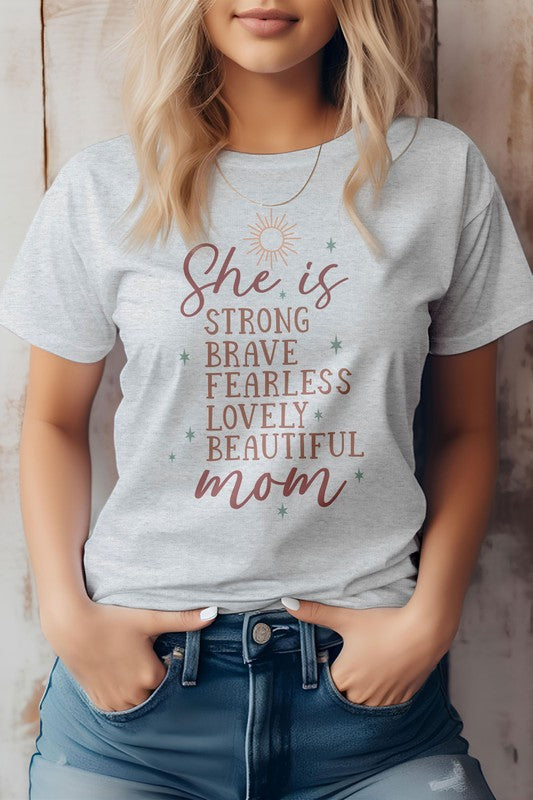 A woman wearing a light pink "She is Mom Graphic Tee" with text that reads, "She is strong, brave, fearless, lovely, beautiful mom." She has blonde hair and her hands are in her jean pockets—a perfect Mother's Day tee.