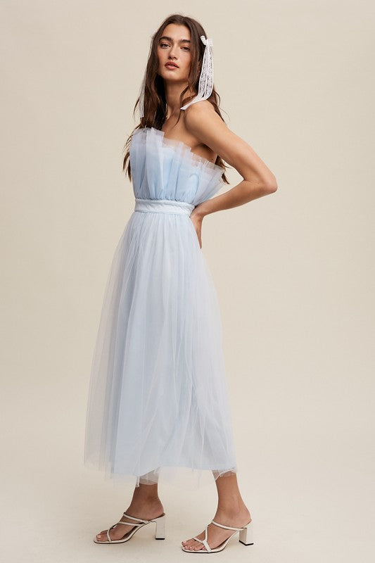 A woman in a light blue Paper Bag Frill Tulle Maxi Dress, featuring a removable adjustable shoulder strap, stands against a plain background in white heels, holding the skirt of her dress with one hand.