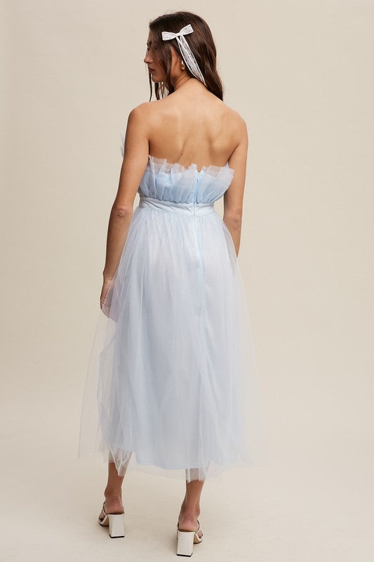 A woman in a light blue Paper Bag Frill Tulle Maxi Dress, featuring a removable adjustable shoulder strap, stands against a plain background in white heels, holding the skirt of her dress with one hand.