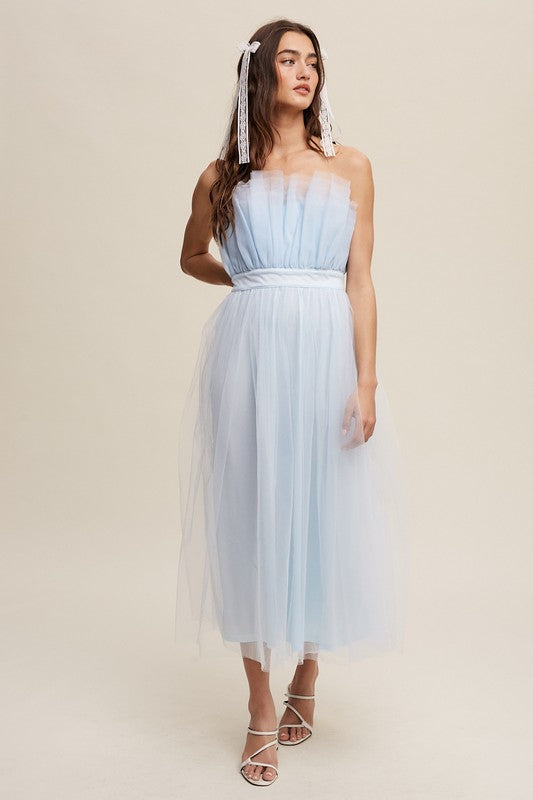 A woman in a light blue Paper Bag Frill Tulle Maxi Dress, featuring a removable adjustable shoulder strap, stands against a plain background in white heels, holding the skirt of her dress with one hand.