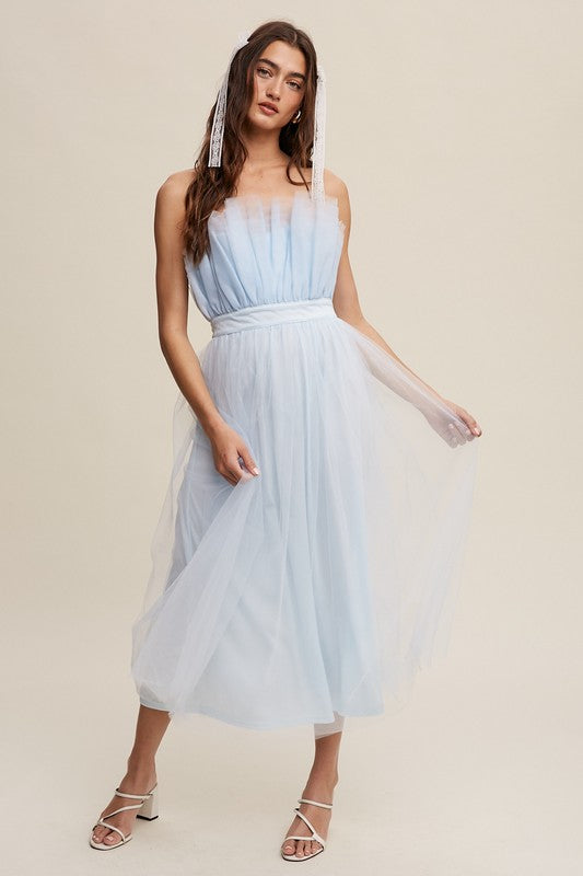 A woman in a light blue Paper Bag Frill Tulle Maxi Dress, featuring a removable adjustable shoulder strap, stands against a plain background in white heels, holding the skirt of her dress with one hand.