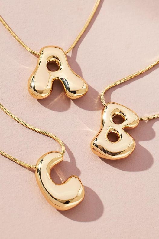 Three Water Drop Bubble Letter Initial Letter Necklaces, each gold-plated and featuring pendant letters "A," "B," and "C," are displayed on a light pink background.