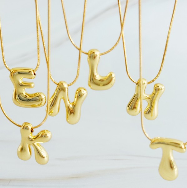 Gold necklaces featuring distinctive, balloon-shaped pendants of the letters E, N, K, L, M, and T against a white backdrop. These Balloon Letter Initial Necklaces are trend-setting and crafted in 18k gold plating for a stylish complement to any ensemble.