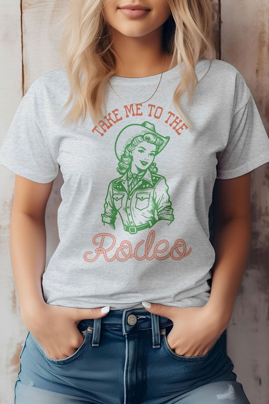 A woman is wearing the "Take Me To The Rodeo, Retro Western Graphic Tee," a light green shirt featuring a vintage-style cowgirl graphic and the text "TAKE ME TO THE RODEO," made from eco-friendly materials, with her hands in jean pockets.