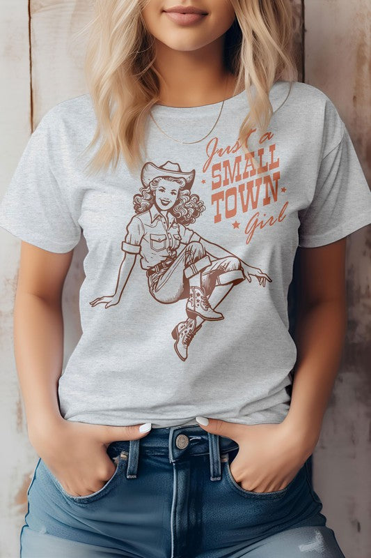 A woman wearing the "Just a Small Town Girl" Retro Western Graphic Tee, which features a beige color, the text "Just a Small Town Girl," and a vintage-style cowgirl illustration. This unisex fit shirt is made from eco-friendly materials, offering both style and sustainability.