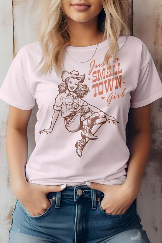 A woman wearing the "Just a Small Town Girl" Retro Western Graphic Tee, which features a beige color, the text "Just a Small Town Girl," and a vintage-style cowgirl illustration. This unisex fit shirt is made from eco-friendly materials, offering both style and sustainability.