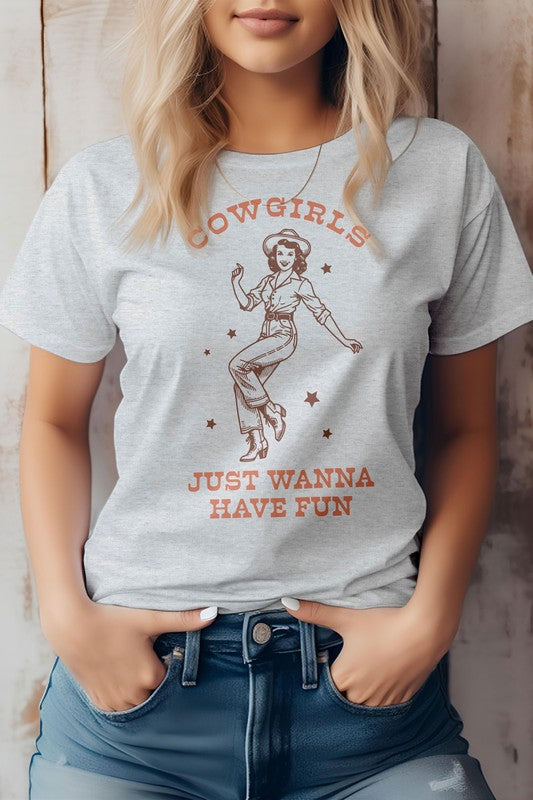 A person wearing the "Cowgirls Just Wanna Have Fun, Western Graphic Tee" featuring a retro illustration of a cowgirl and the caption "Cowgirls just wanna have fun," standing against a neutral background.