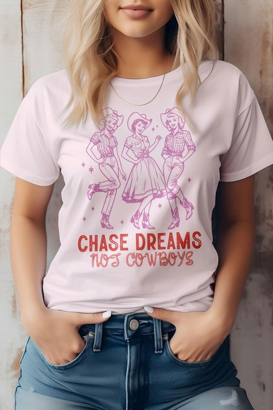 A person wearing the Chase Dreams Not Cowboys, Western Graphic Tee showcases a design of three line-dancing figures and the phrase "CHASE DREAMS NOT COWBOYS" written below, offering a humor-infused touch to this eco-friendly white t-shirt.