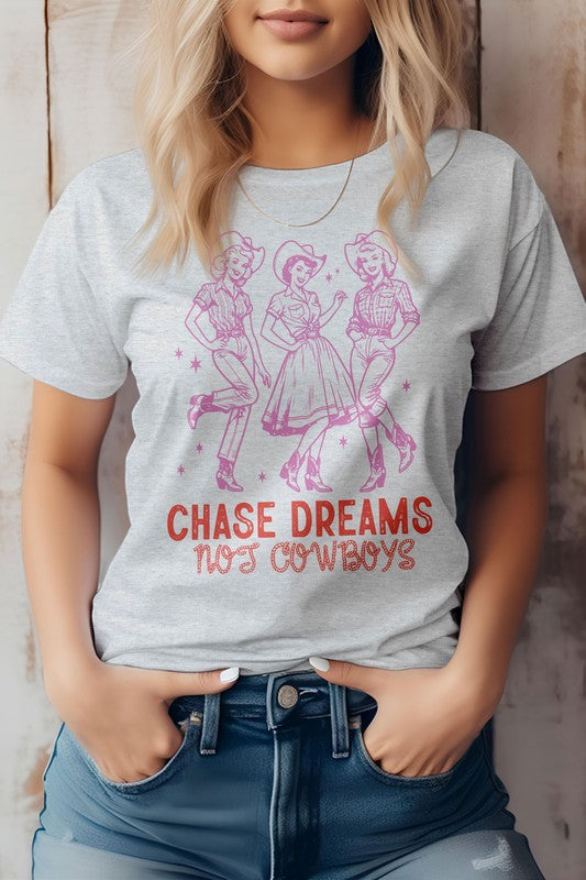 A person wearing the Chase Dreams Not Cowboys, Western Graphic Tee showcases a design of three line-dancing figures and the phrase "CHASE DREAMS NOT COWBOYS" written below, offering a humor-infused touch to this eco-friendly white t-shirt.