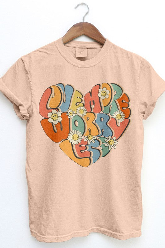 The Love More Worry Less Retro Garment Dye Tee, a beige T-shirt with a vibrant heart graphic and the phrase "LOVE MORE WORRY LESS," adorned with daisy flowers, hangs on a wooden hanger. This unisex graphic tee embodies a retro streetwear vibe, making it a timeless addition to your wardrobe.