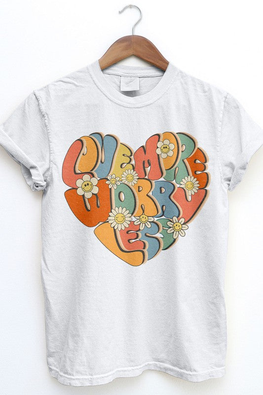 The Love More Worry Less Retro Garment Dye Tee, a beige T-shirt with a vibrant heart graphic and the phrase "LOVE MORE WORRY LESS," adorned with daisy flowers, hangs on a wooden hanger. This unisex graphic tee embodies a retro streetwear vibe, making it a timeless addition to your wardrobe.