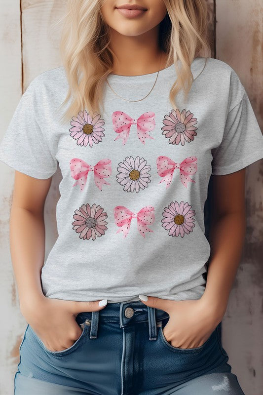 A person wearing a light pink Coquette Pink Bow Daisy Flower Boho Graphic Tee, which showcases alternating flowers and pink bows. They have their hands in the pockets of their blue jeans.