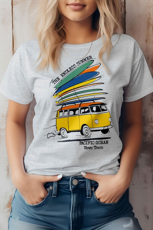 A woman is wearing The Endless Summer Graphic Tee, which features a vintage design with a yellow van and surfboards, alongside the text "The Endless Summer, Pacific Ocean, Wave Team." With her hands in her jeans pockets, she exudes California surf vibes.