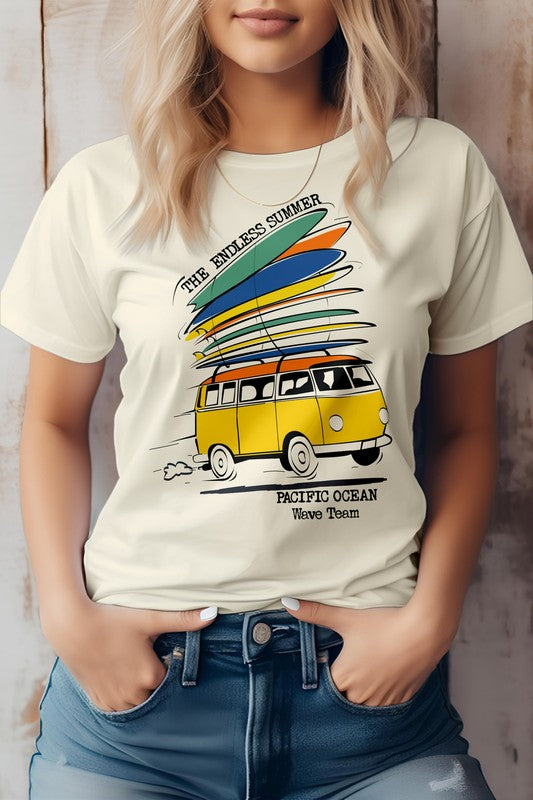 A woman is wearing The Endless Summer Graphic Tee, which features a vintage design with a yellow van and surfboards, alongside the text "The Endless Summer, Pacific Ocean, Wave Team." With her hands in her jeans pockets, she exudes California surf vibes.