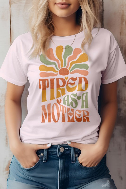 A person wearing the Tired as a Mother Retro Graphic Tee, featuring colorful retro text that reads "TIRED AS A MOTHER" alongside a design resembling a sunrise in beige. Their hands are in the pockets of their jeans, perfectly capturing the essence of Mom Life.