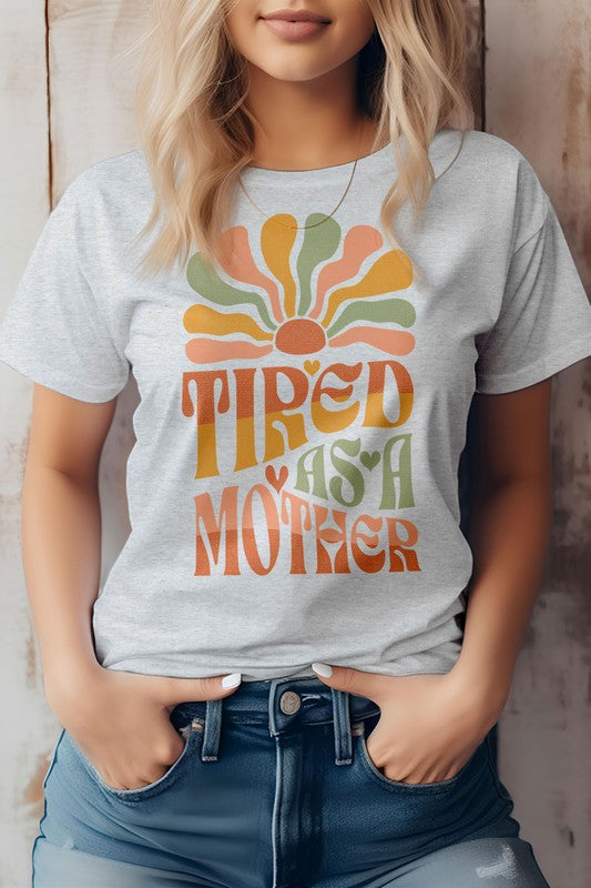 A person wearing the Tired as a Mother Retro Graphic Tee, featuring colorful retro text that reads "TIRED AS A MOTHER" alongside a design resembling a sunrise in beige. Their hands are in the pockets of their jeans, perfectly capturing the essence of Mom Life.