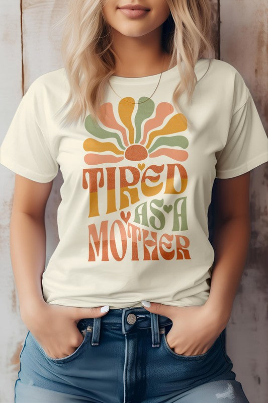 A person wearing the Tired as a Mother Retro Graphic Tee, featuring colorful retro text that reads "TIRED AS A MOTHER" alongside a design resembling a sunrise in beige. Their hands are in the pockets of their jeans, perfectly capturing the essence of Mom Life.