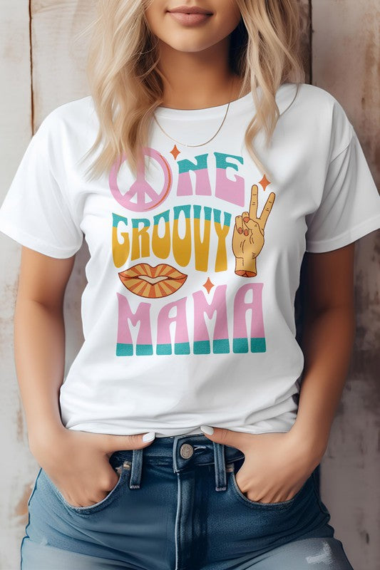 A person wearing a light gray "One Groovy Mama" Retro Quote Hippie Graphic Tee, featuring a peace sign, a hand making a peace gesture, and a pair of lips. The shirt, crafted from superior combed and ring-spun cotton, complements the person's blonde hair as they keep their hands in their pockets.