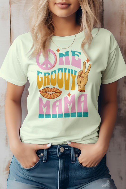 A person wearing a light gray "One Groovy Mama" Retro Quote Hippie Graphic Tee, featuring a peace sign, a hand making a peace gesture, and a pair of lips. The shirt, crafted from superior combed and ring-spun cotton, complements the person's blonde hair as they keep their hands in their pockets.