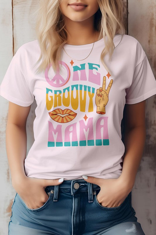 A person wearing a light gray "One Groovy Mama" Retro Quote Hippie Graphic Tee, featuring a peace sign, a hand making a peace gesture, and a pair of lips. The shirt, crafted from superior combed and ring-spun cotton, complements the person's blonde hair as they keep their hands in their pockets.